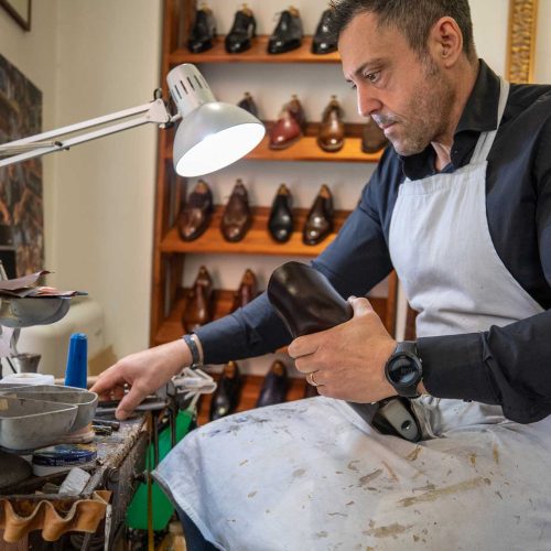 Giò seduto al banco tiene una scarpa sulla forma e cerca un attrezzo